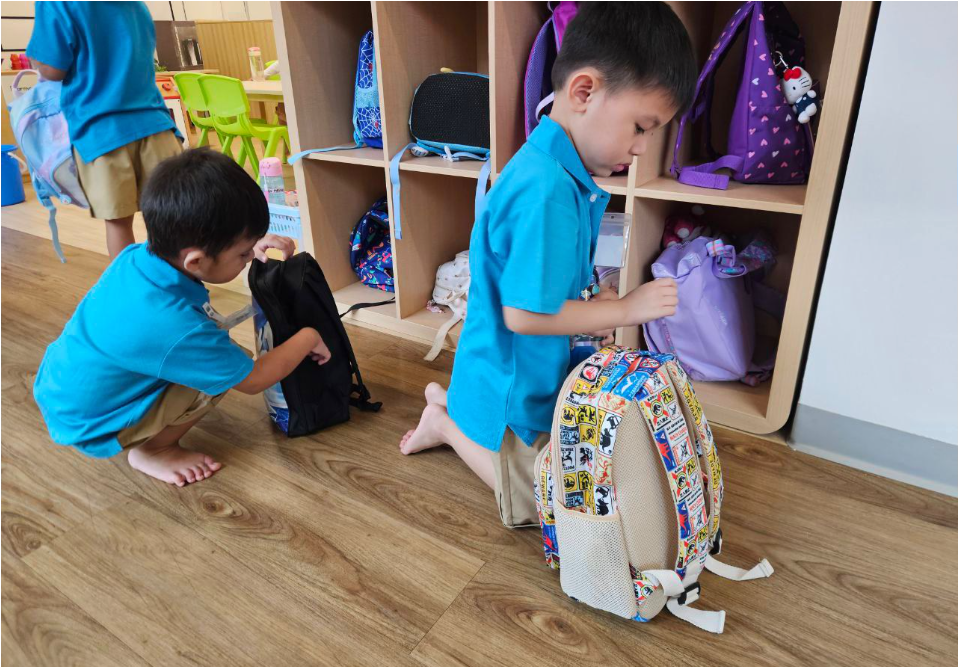 Two children in blue shirts kneel by cubbies, packing backpacks in a classroom.
